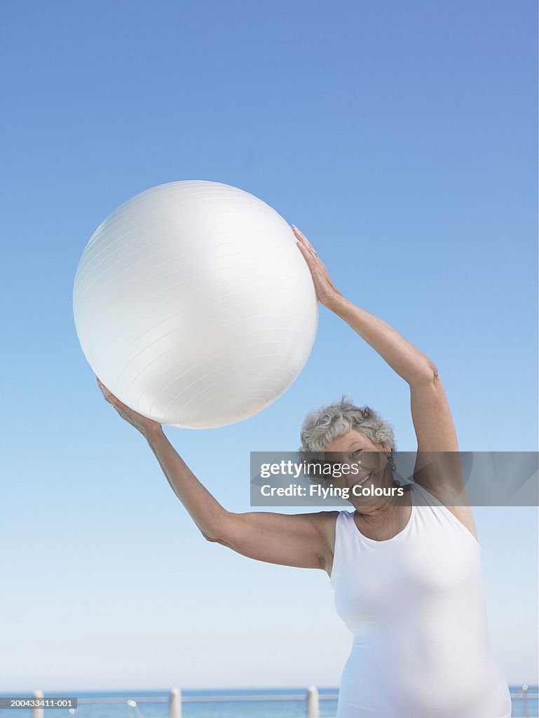 Senior woman holding exercise ball over head, smiling, portrait