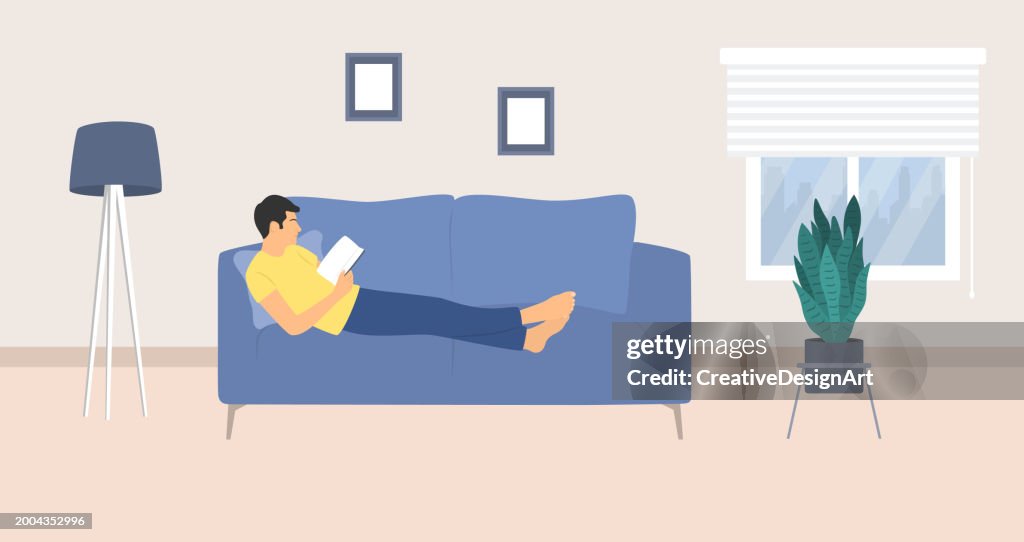 Side View Of Relaxed Young Man Reading Book On Couch. Living Room Interior With Floor Lamp, Sofa And Potted Plant