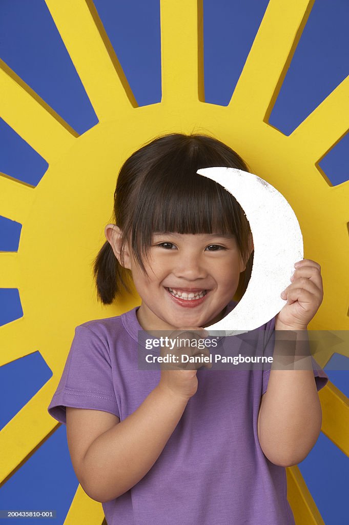 Girl (2-4) holding paper moon by picture of sun, smiling, portrait