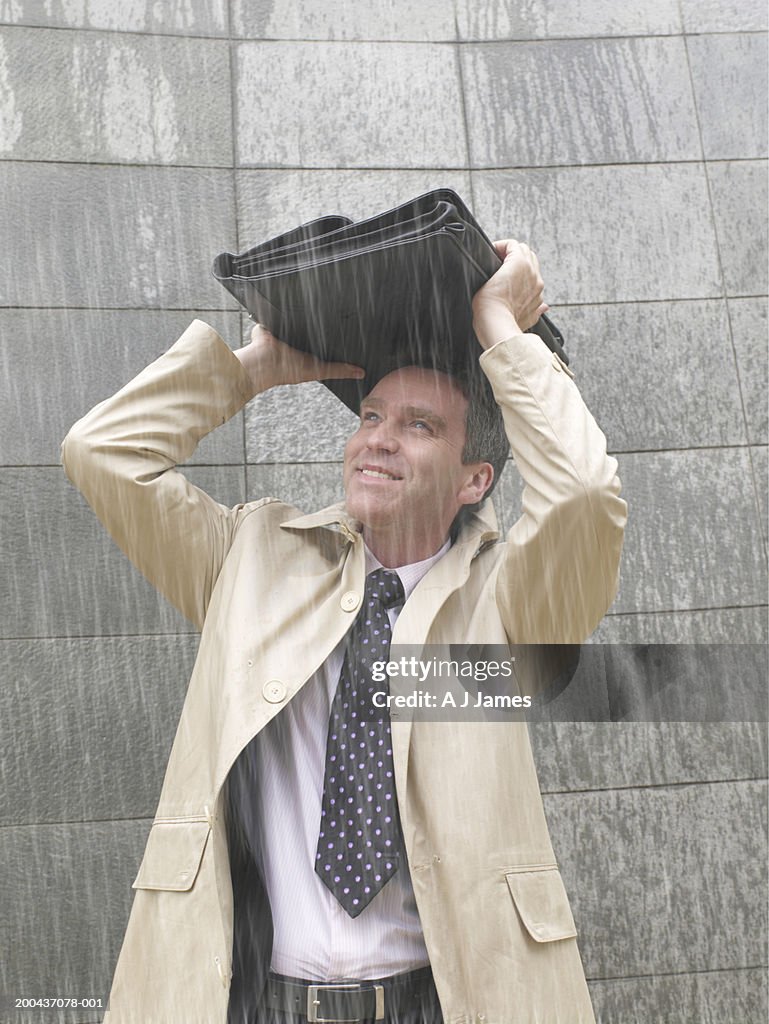 Businessman standing in rain holding briefcase over head, smiling