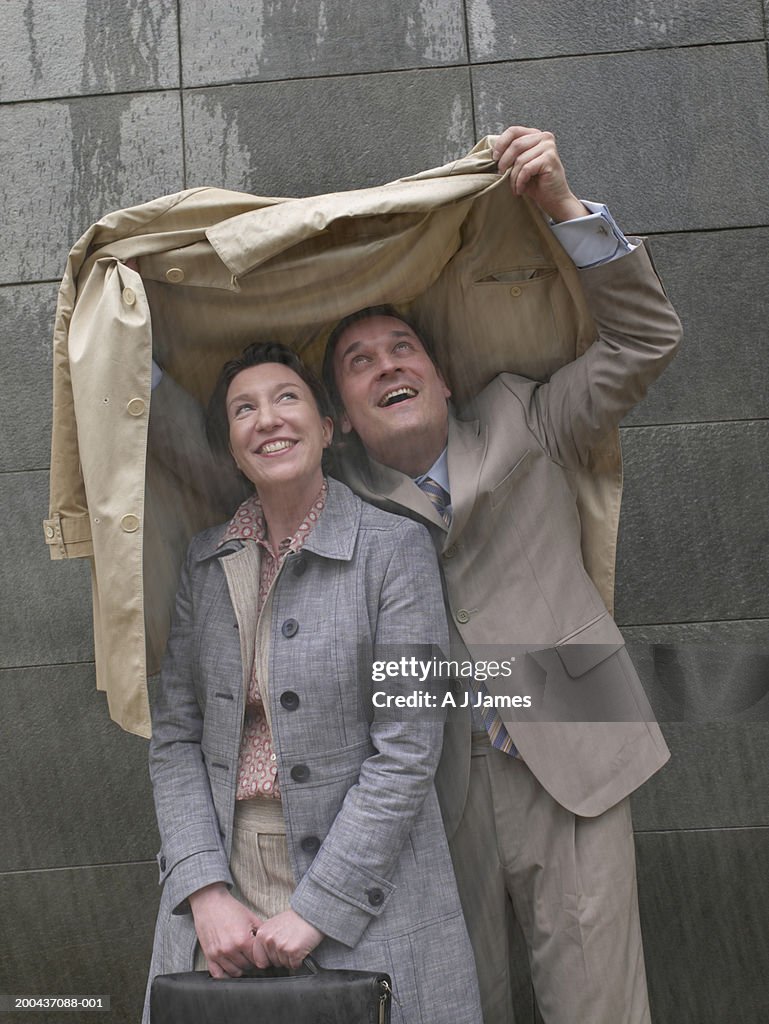 Businessman and woman sheltering under man's coat in rain, smiling