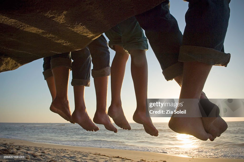 Parents and children dangling feet from ledge above beach, low section, sunset