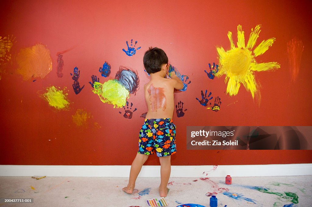 Boy (3-5) fingerpainting on red wall, rear view
