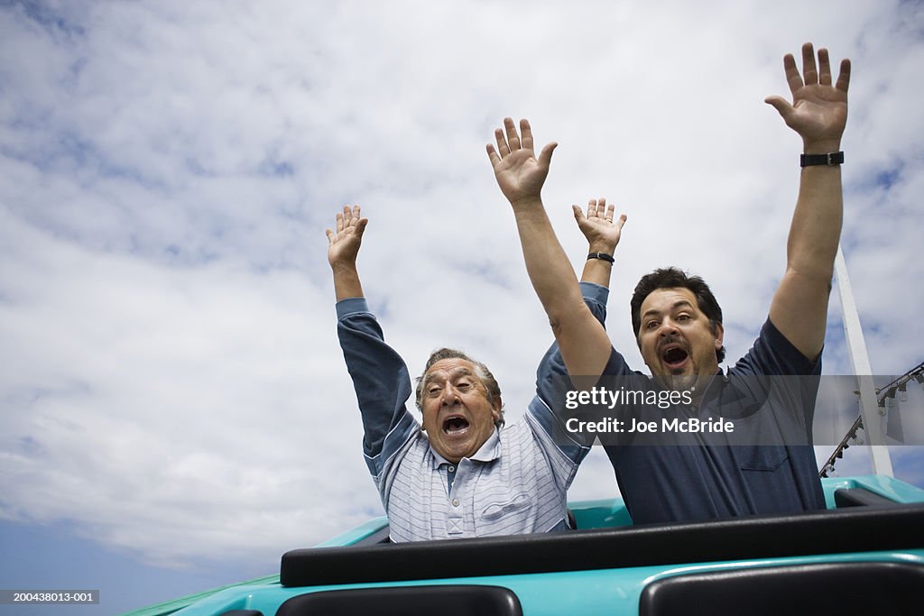 Father and adult son riding rollercoaster, hands in air