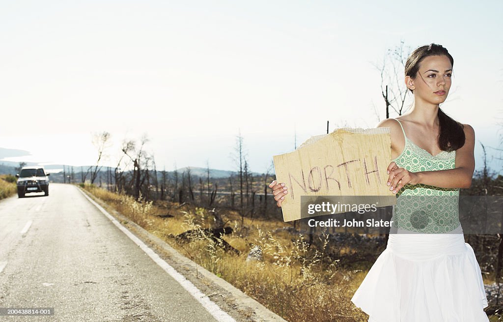 Young woman hitchhiking at side of road, holding up 'North' sign