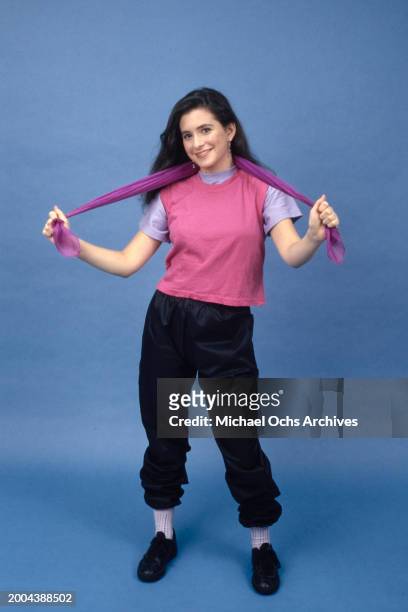 Model wearing a pink singlet over a lilac t-shirt with black trousers tucked into her socks, holding a purple scarf, against a blue background, in a...