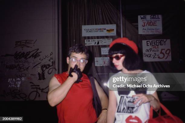 Two people, one wearing a sleeveless red t-shirt and black gloves, the other wearing a t-shirt featuring a pair of sunglasses above a pair of lips, a...