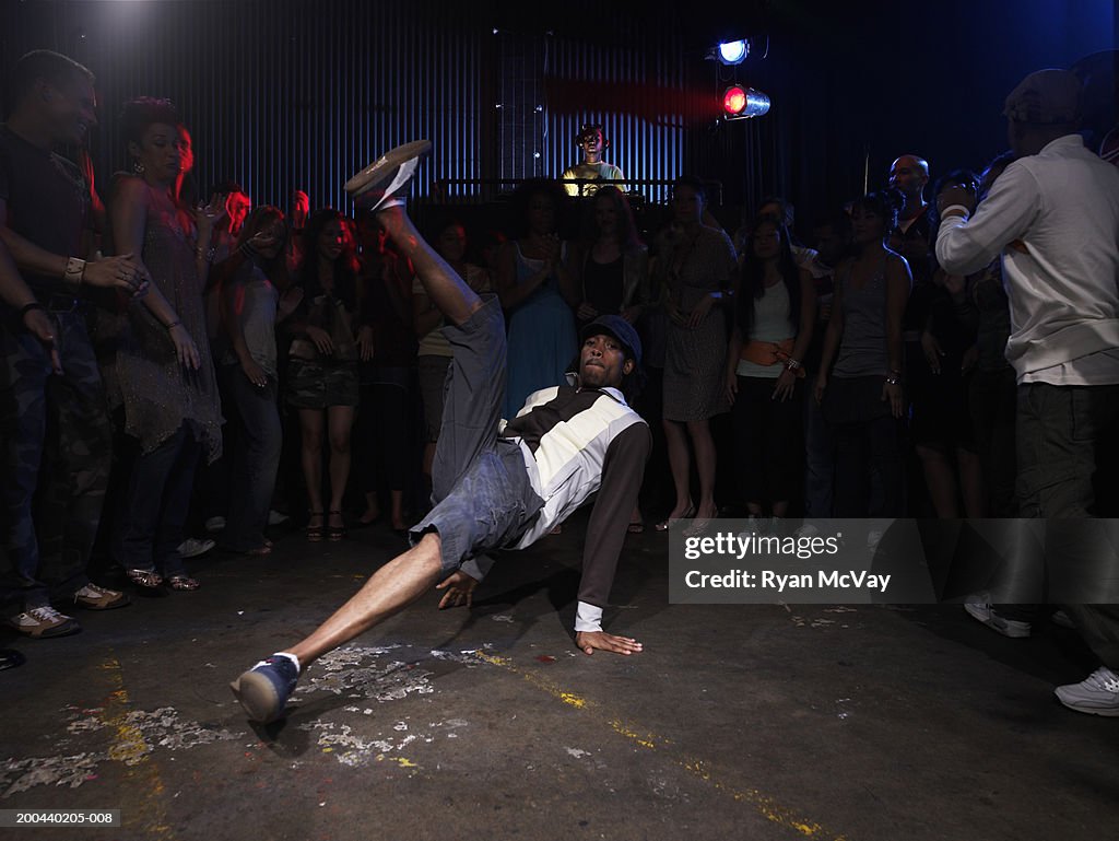 Group of adults watching man breakdance in club, DJ in background