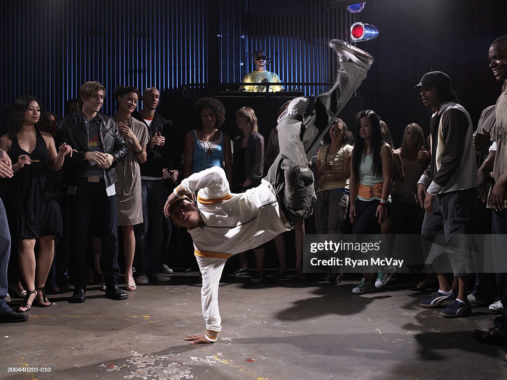 Group of adults watching man breakdance in club, DJ in background