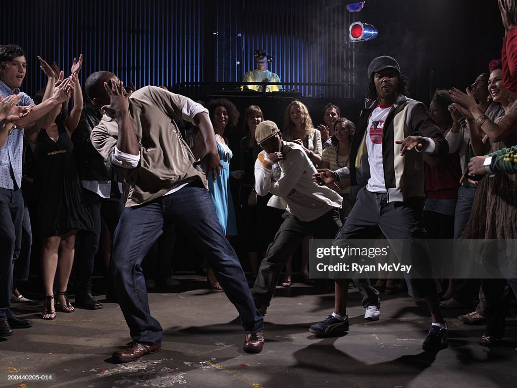 Crowd watching three men breakdance in club, DJ in background