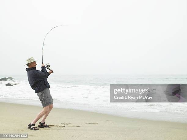 mature man reeling in catch on beach - fishing reel foto e immagini stock