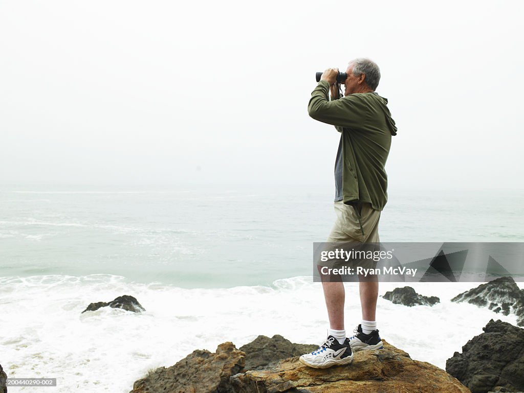 Mature man with binoculars standing on rocks on beach