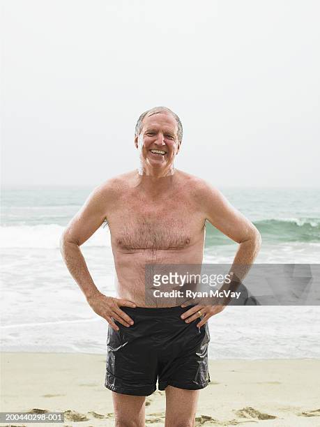 mature man in shorts standing on beach, hands on hips - swimming shorts stock pictures, royalty-free photos & images