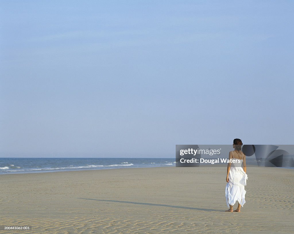 Young woman standing on beach wearing white dress, rear view
