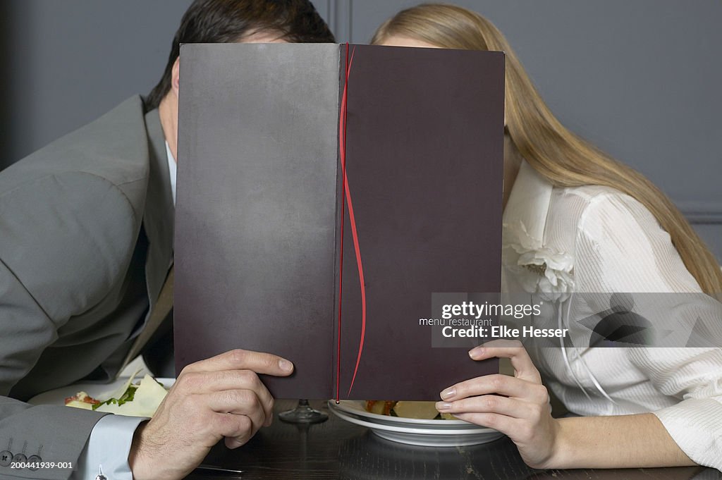 Couple at restaurant table leaning towards each other behind menu