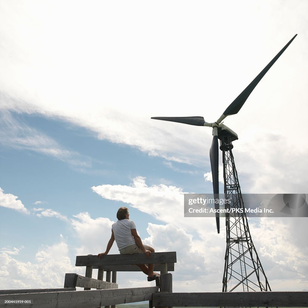 Man on corral gate watching wind turbine, rear view