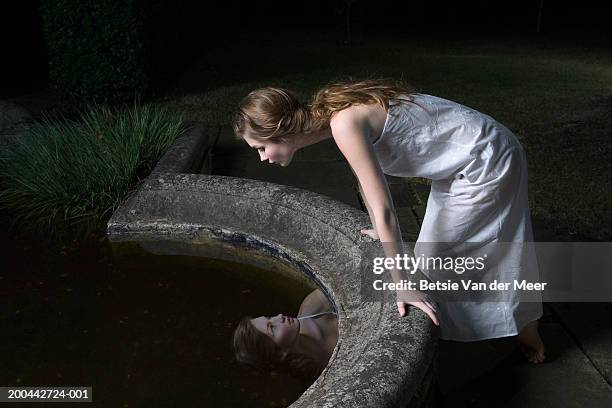 teenage girl (16-18) leaning over reflection in garden pond, night - eitelkeit stock-fotos und bilder