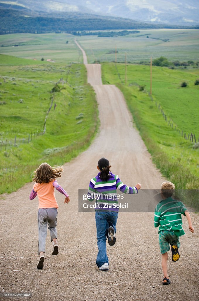 Three children (8-12) running on country road, rear view