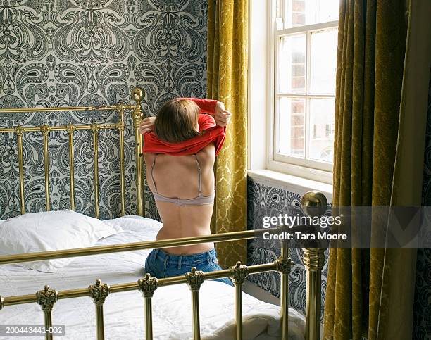 young woman removing top sitting on bed by window, rear view - spogliarsi foto e immagini stock