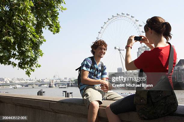 england, london, couple sitting on embankment, woman photographing man - london eye stockfoto's en -beelden