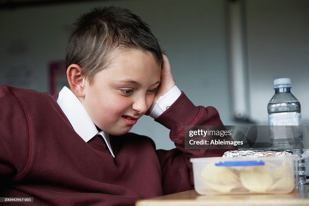 Schoolboy (8-10) sitting at desk staring at lunchbox, pulling face