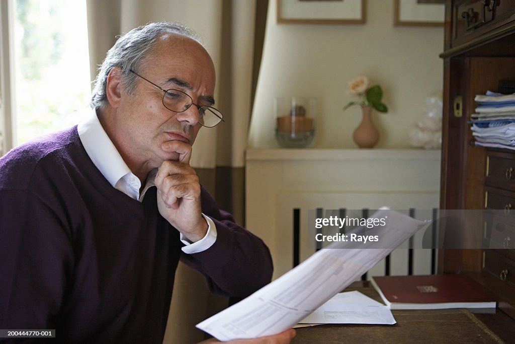 Mature man sitting in study looking at bills, frowning