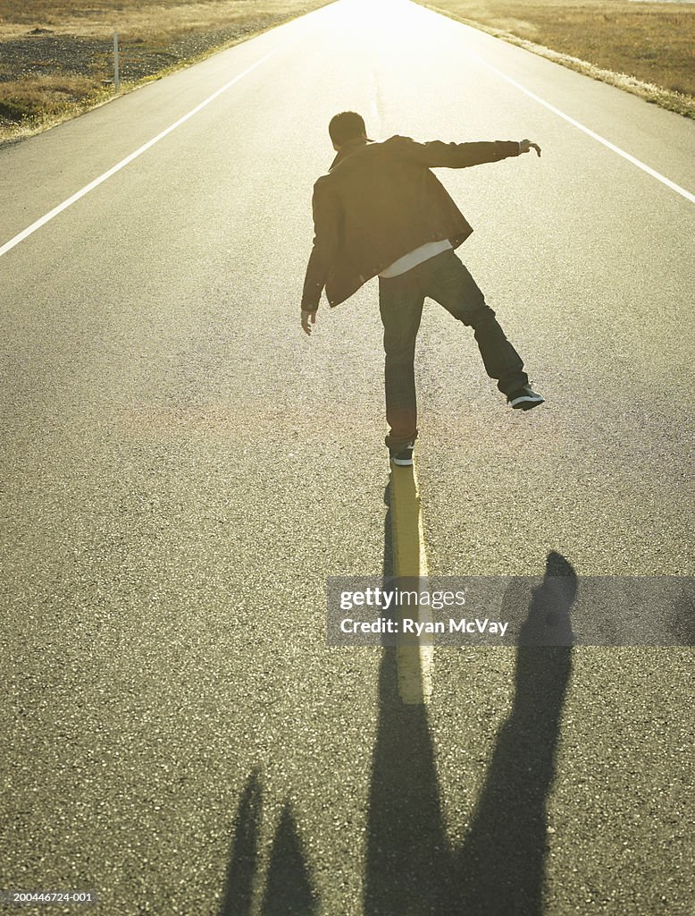Young man walking along line in middle of road at sunset, rear view