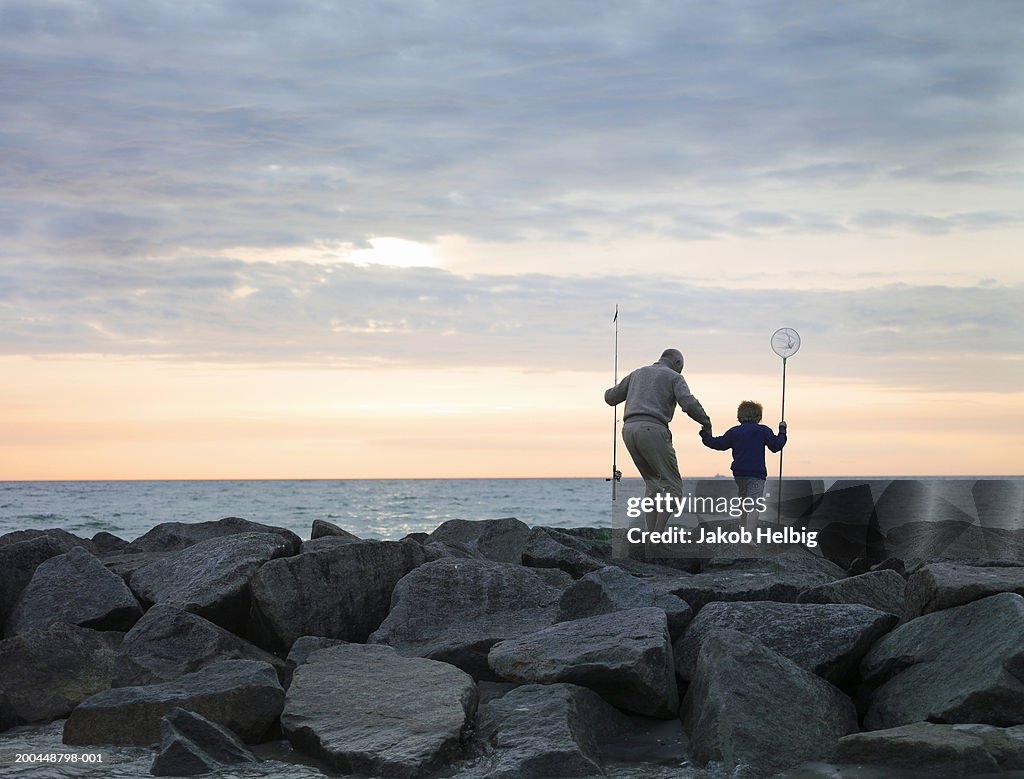 Grandfather and grandson (4-6) walking on rocks with fishing equipment