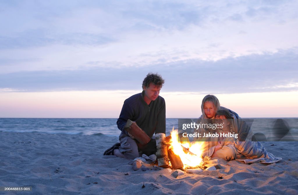 Parents and daughter (6-8)sitting around fire on beach, sunset