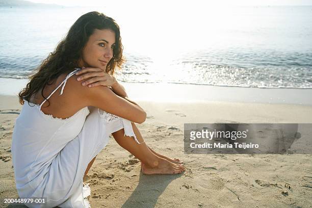 young woman wearing white dress, sitting on beach - wavy hair beach stock pictures, royalty-free photos & images