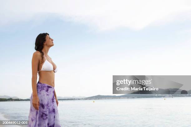 young woman wearing bikini top and sarong, standing on beach - sarong stock pictures, royalty-free photos & images