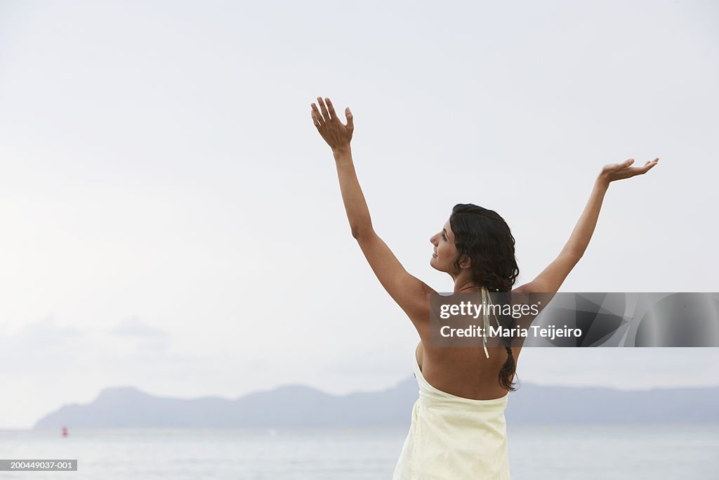 Young woman standing on beach, arms raised, rear view
