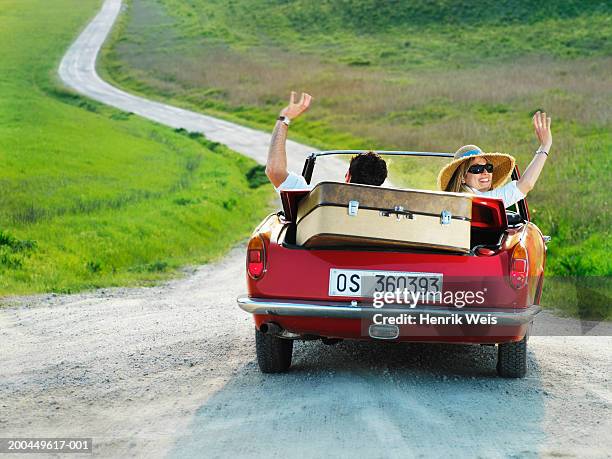 Couple Driving Convertible Car In Countryside Waving Arms Rear