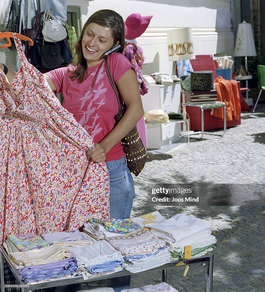 Woman using mobile phone, looking at dress at outdoor market, smiling