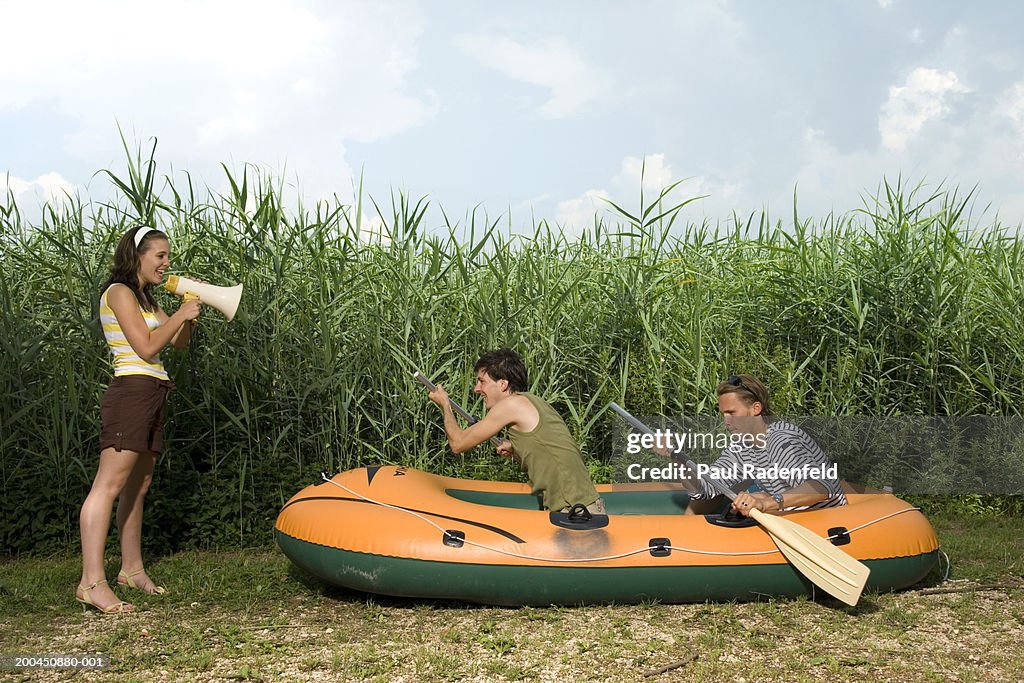 Men in dinghy rowing on dryland with young woman using megaphone