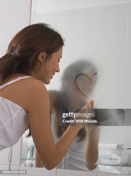 young woman drawing heart shape in foggy bathroom mirror - mirror steam stockfoto's en -beelden