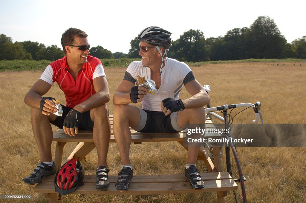 Two male cyclists sitting on picnic table, eating