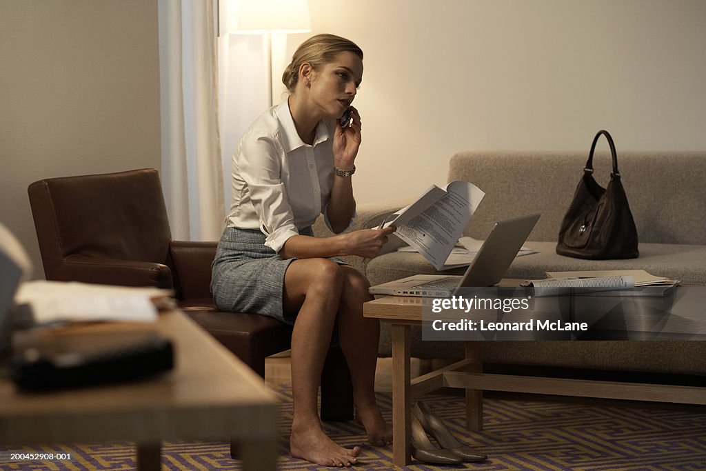 Businesswoman in hotel room holding paperwork while using phone