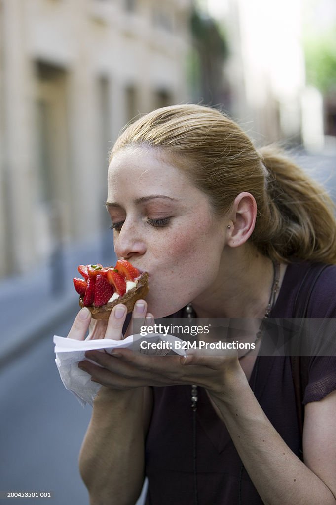 Young woman eating strawberry-topped cake, outdoors, close-up