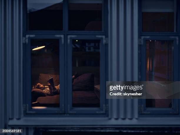 little boy reading a book while relaxing in the evening at home. - photographed through window stockfoto's en -beelden