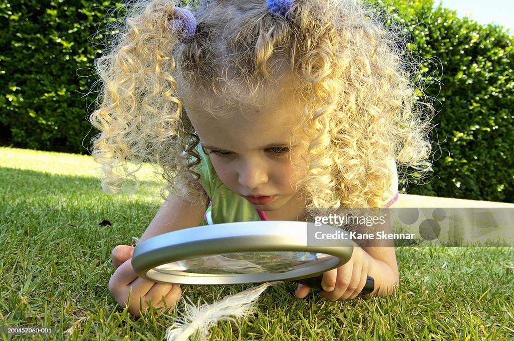 Girl (3-5) lying on grass, looking at feather through magnifying glass