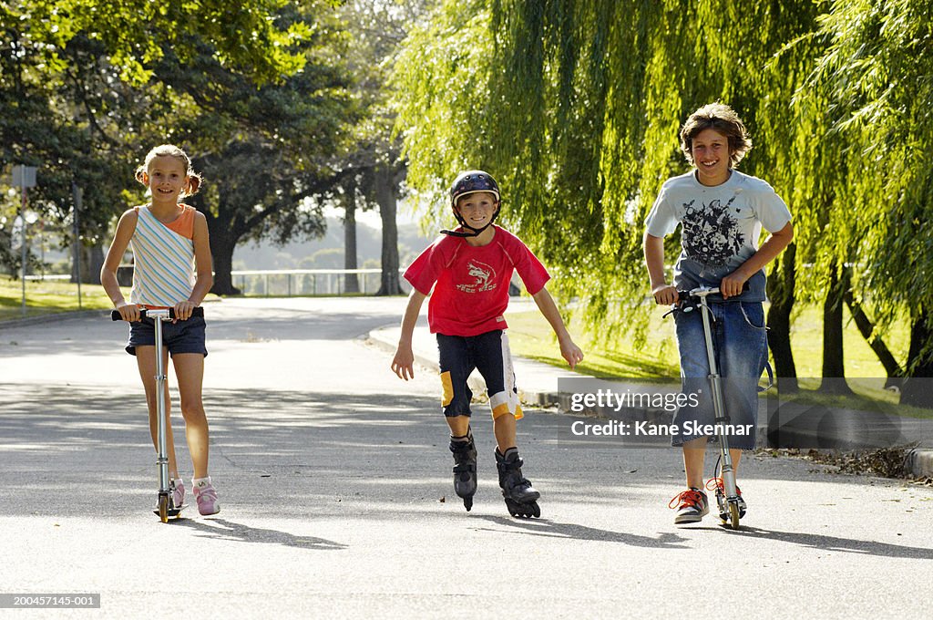 Two boys and girl (9-12)in park,riding scooters and one inline skating