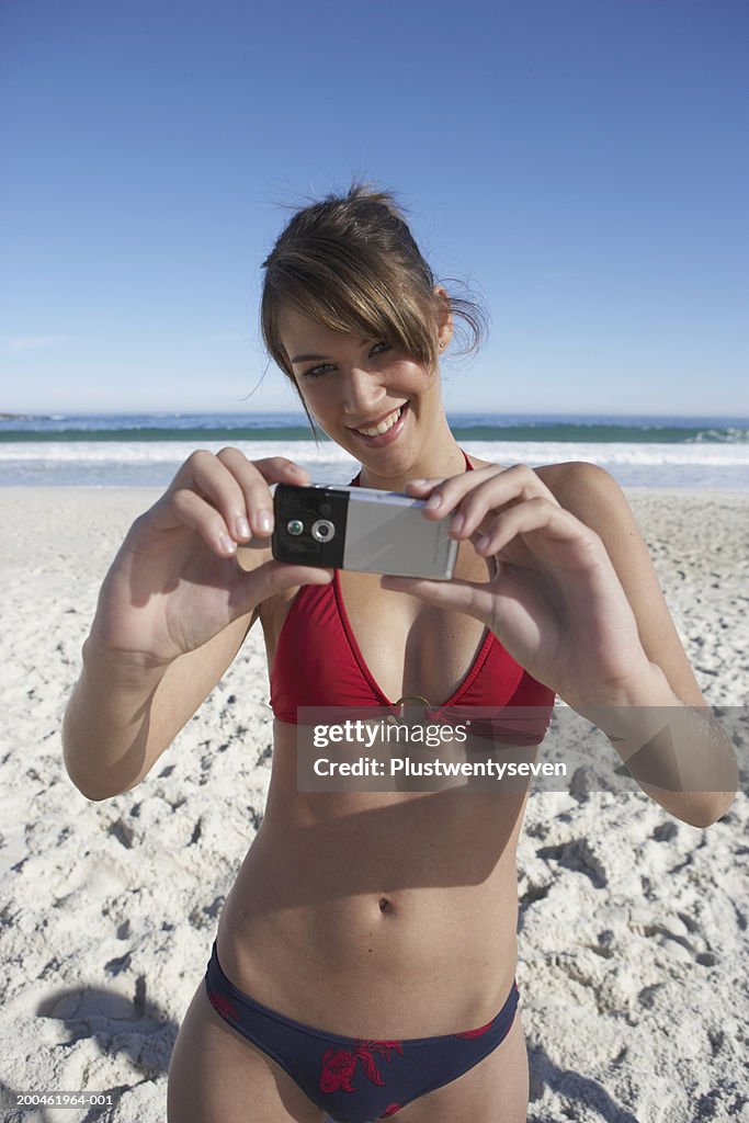 Young woman using camera phone on beach, smiling, portrait