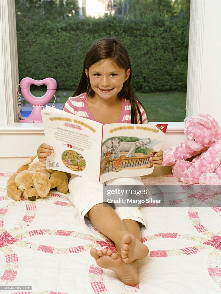 Girl (6-8) sitting on bed with book and stuffed toys, portrait