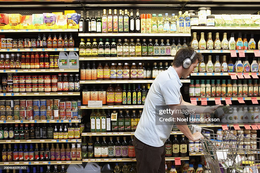 Young man in headphones placing bottle of oil in cart, side view