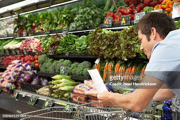 young man reading shopping list in produce aisle, side view, close-up - einkaufszettel stock-fotos und bilder