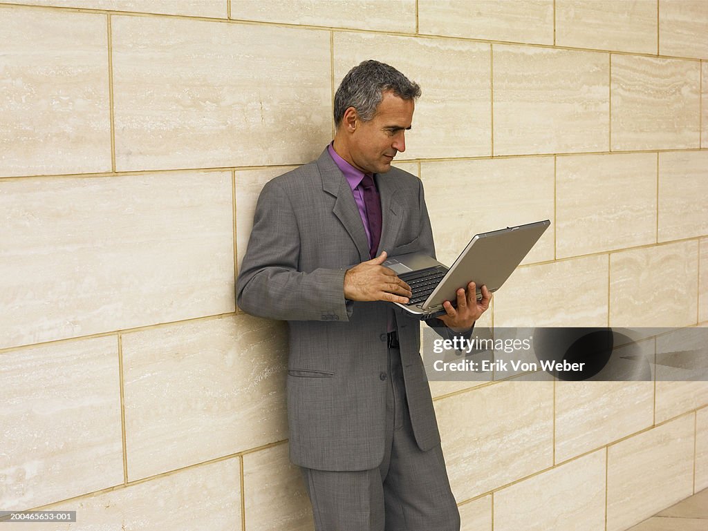 Businessman leaning against wall, using laptop