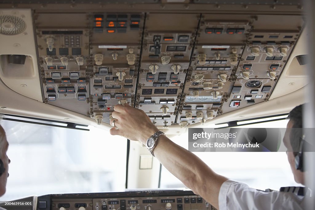 Two pilots in cockpit, one touching control panel, rear view