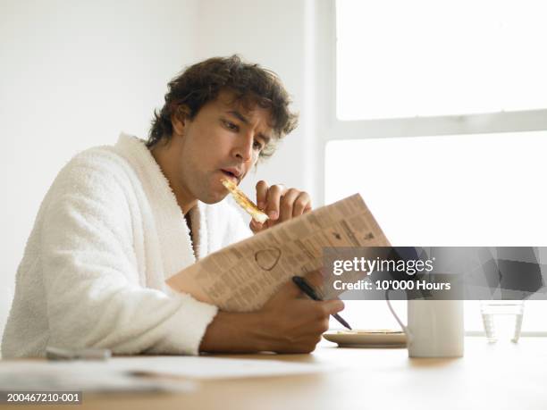 young man in dressing gown reading newspaper whilst eating breakfast - paper gown stock pictures, royalty-free photos & images