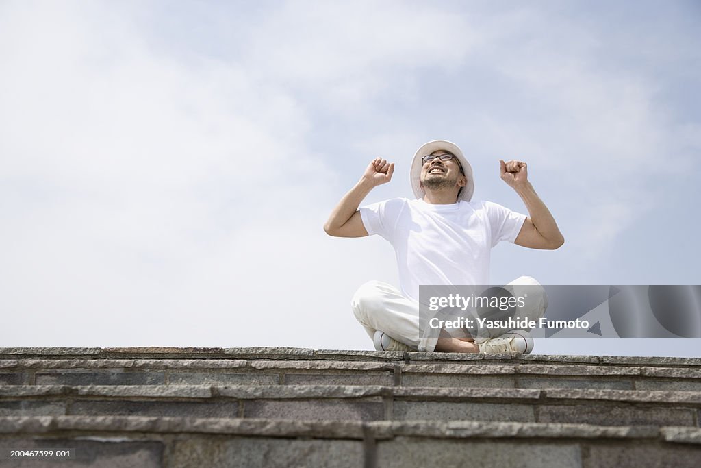 Mature man sitting atop stone wall, stretching and looking up